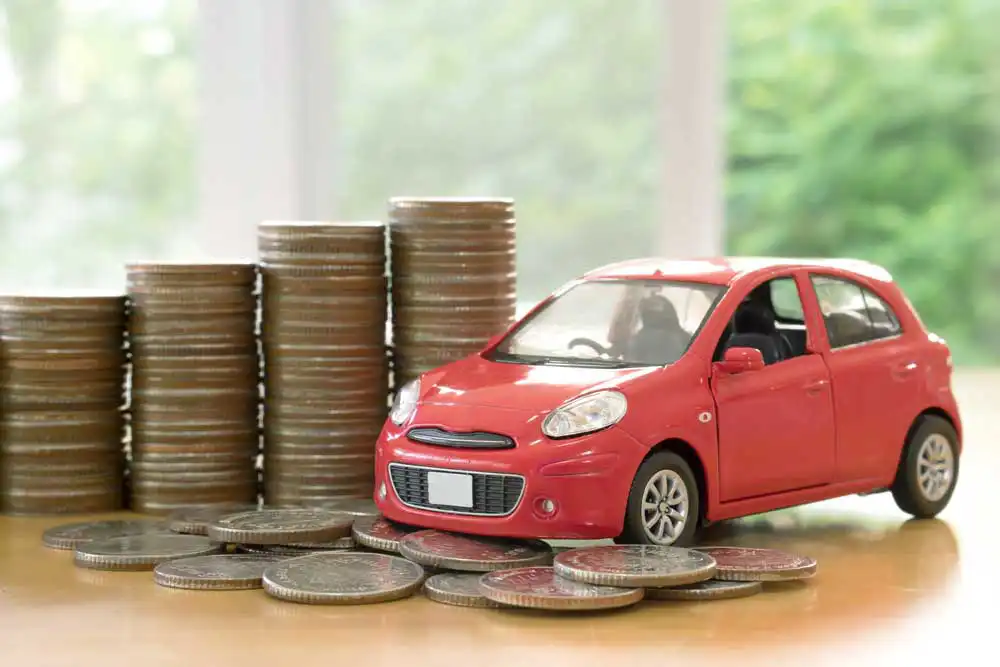 A red, toy, hatchback car with it's front wheels over a pile of coins, with four stacks of coins in the background